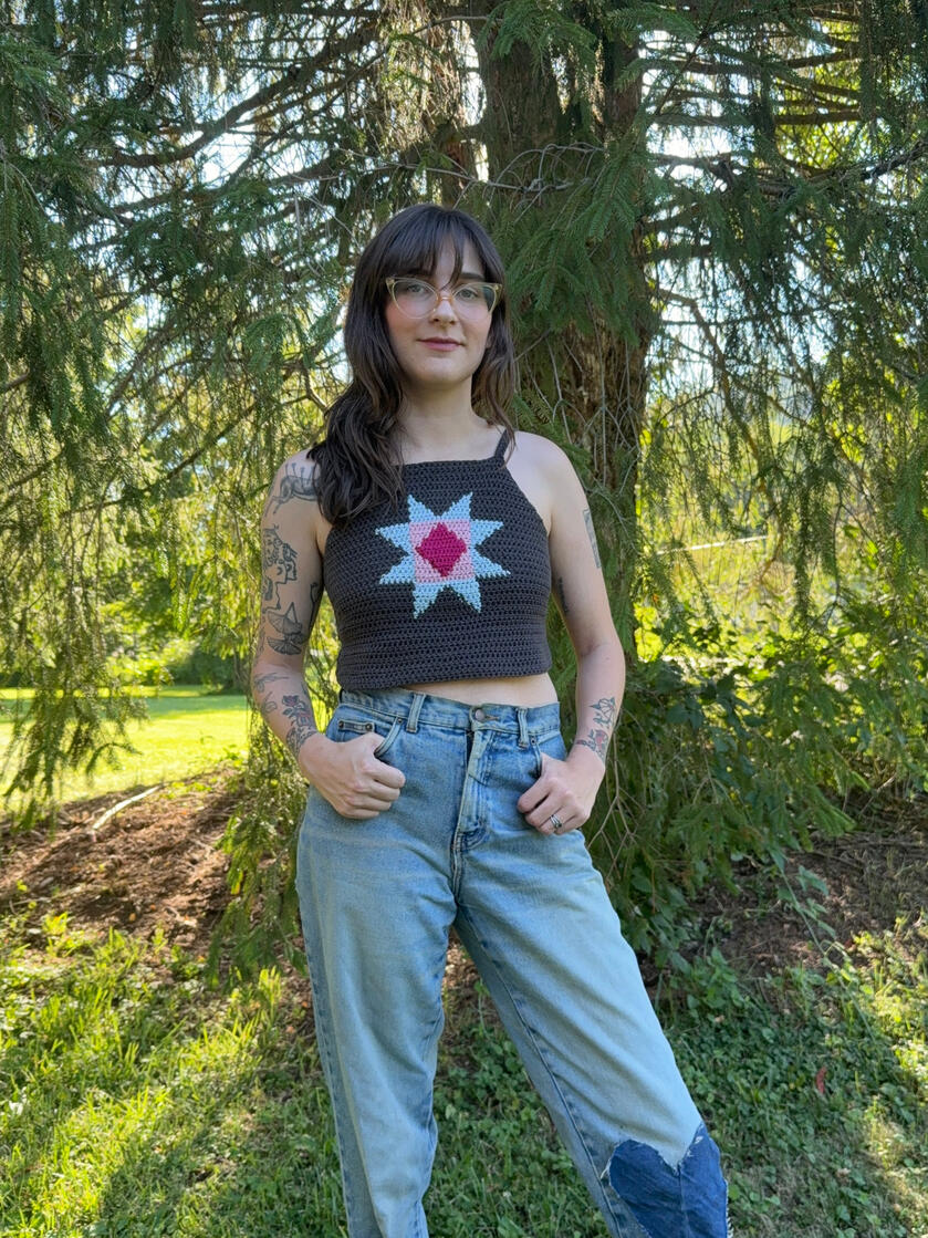 Hannah, with long brown hair and cat eye glasses, wears a quilt inspired crochet top and jeans